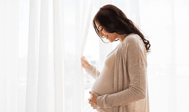 A pregnant woman standing by a bright window, holding her bump